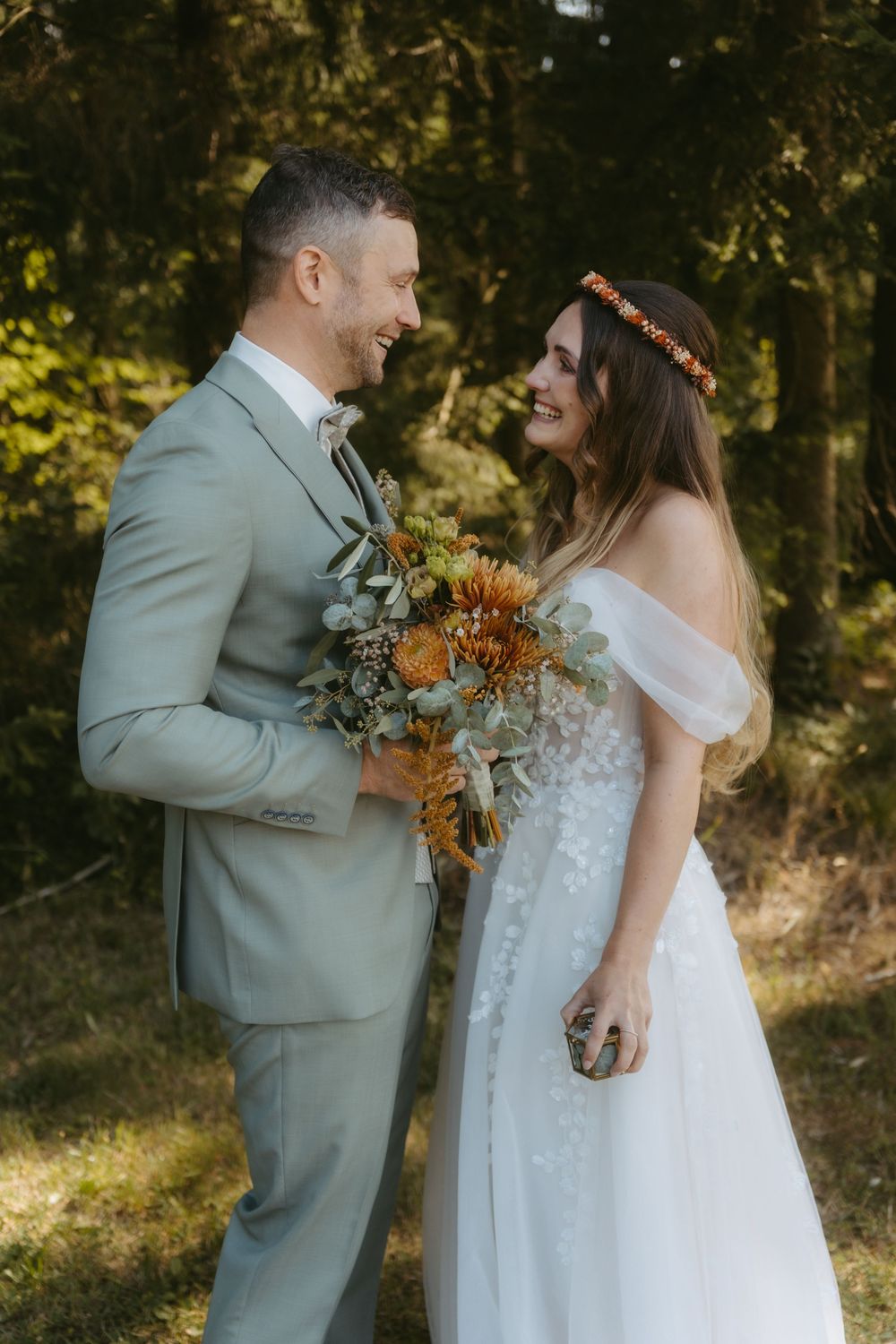 Bride and groom sharing a joyful moment in a forest, holding an autumn orange wedding bouquet.