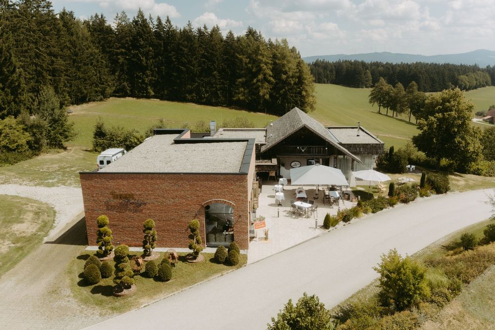 Aerial view of rustic brick wedding venue surrounded by green hills and forest in countryside.
