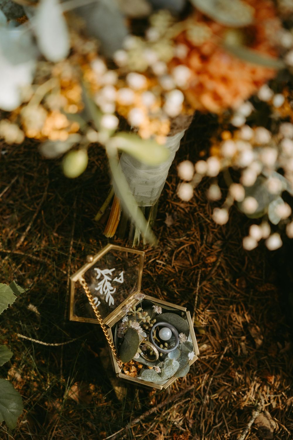 Wedding rings displayed in an open hexagonal glass box surrounded by floral arrangements on ground.