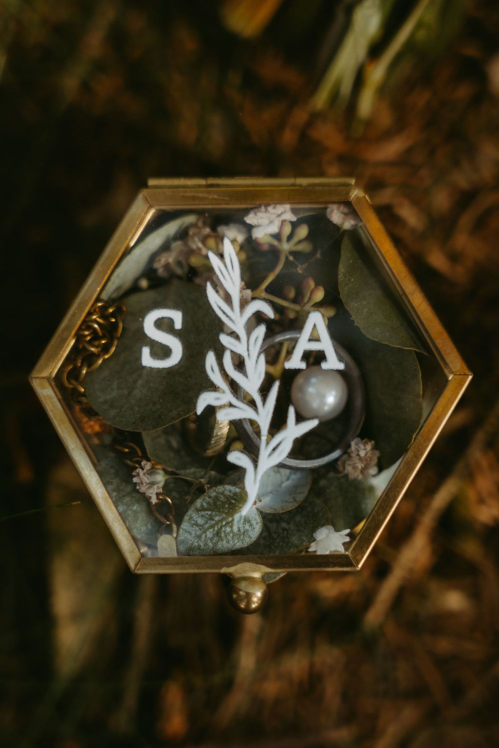 Hexagonal gold glass ring box with initials S and A decorated with botanical elements and wedding rings.