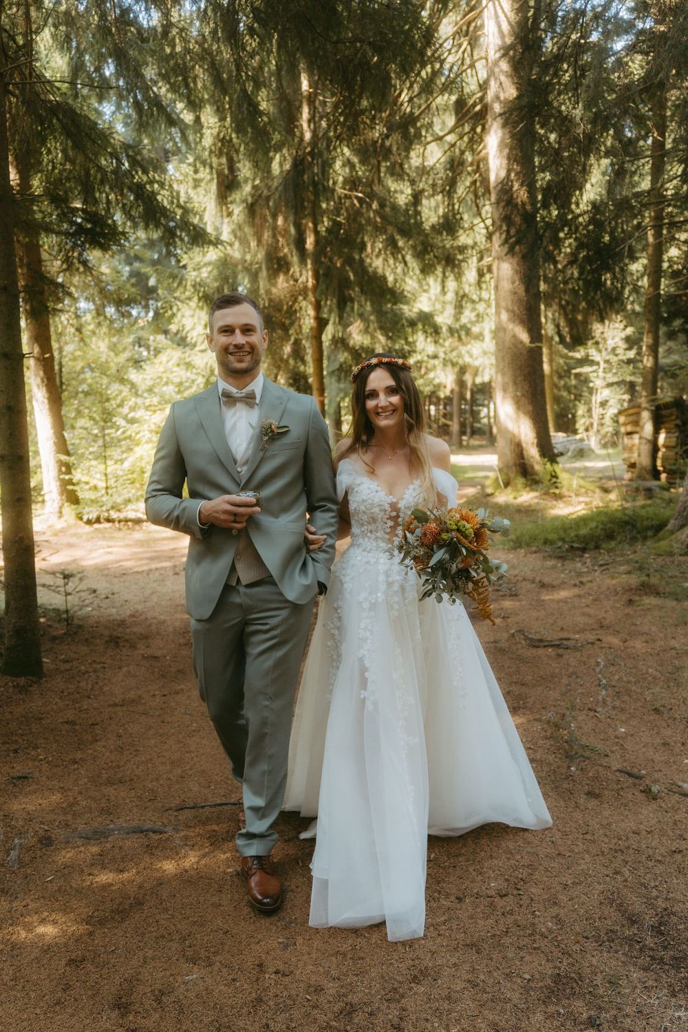 Smiling bride and groom walk hand in hand through a sunlit forest, bride holding a floral bouquet.