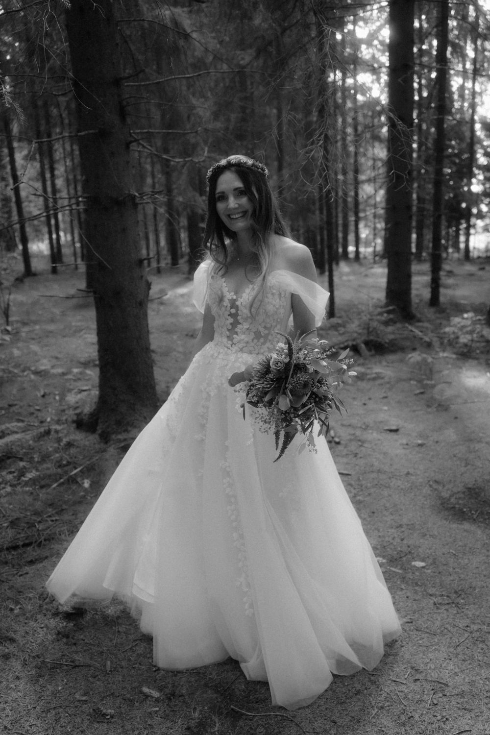 Bride in off-shoulder lace ball gown holding bouquet poses in forest, black and white photo.