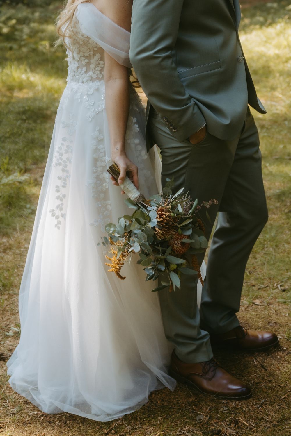 Bride in lace wedding gown holding wildflower bouquet standing beside groom in sage green suit.