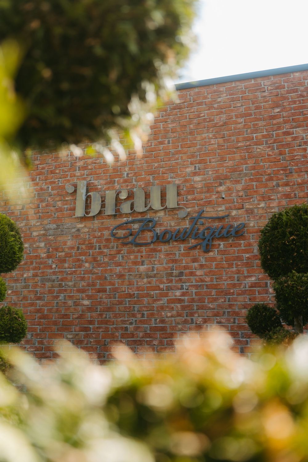 Brick wall exterior of Brau Boutique shop framed by lush green topiary bushes.