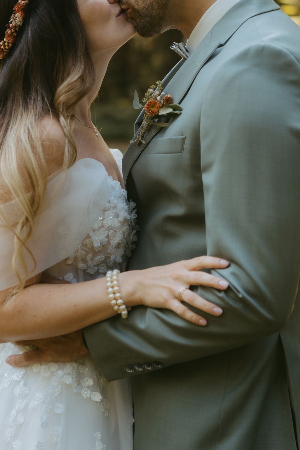 Bride in floral wedding dress with pearl bracelet embraces groom in sage green suit with boutonniere.