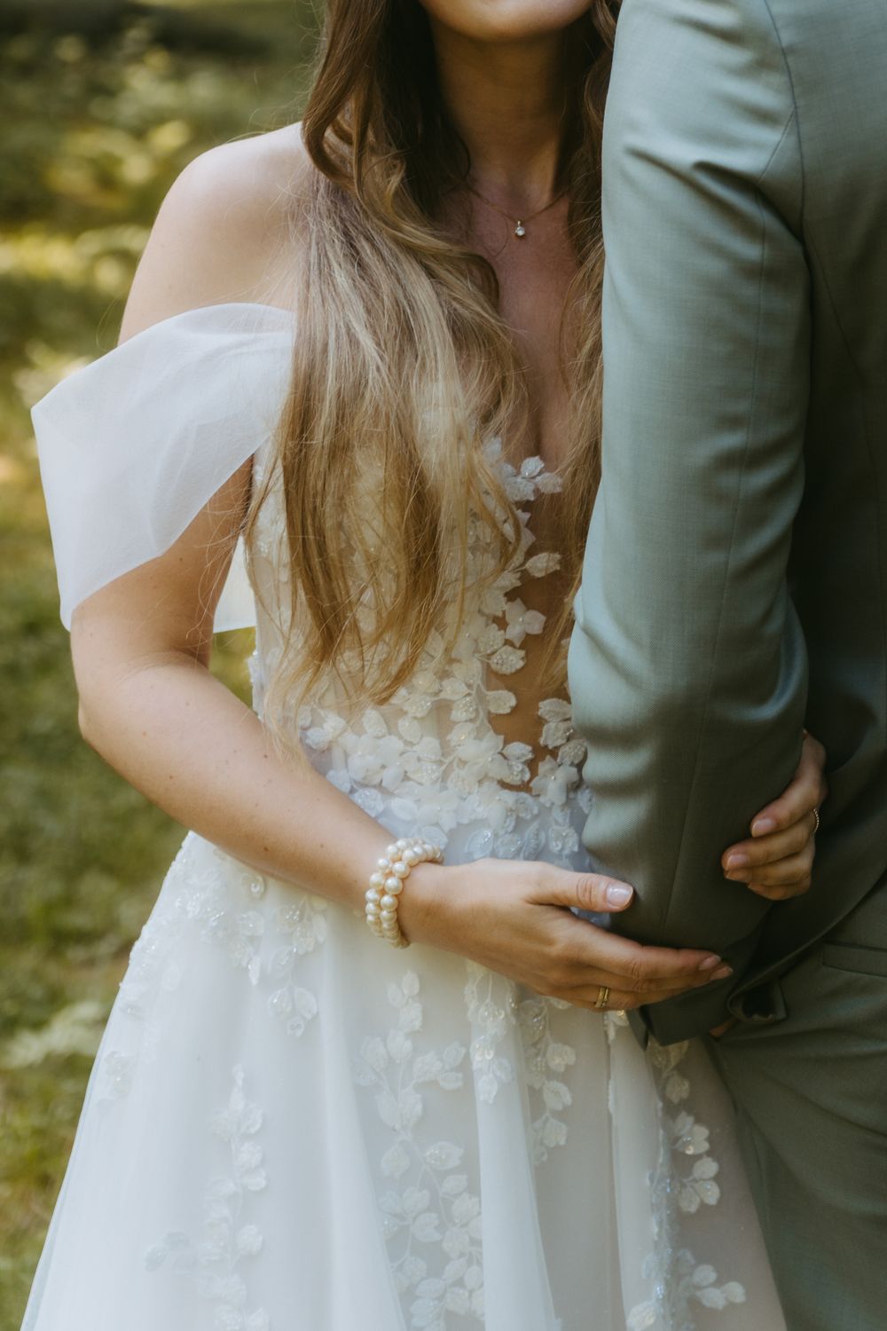 Bride in floral off-shoulder wedding gown with pearl bracelet embracing groom in gray suit outdoors.