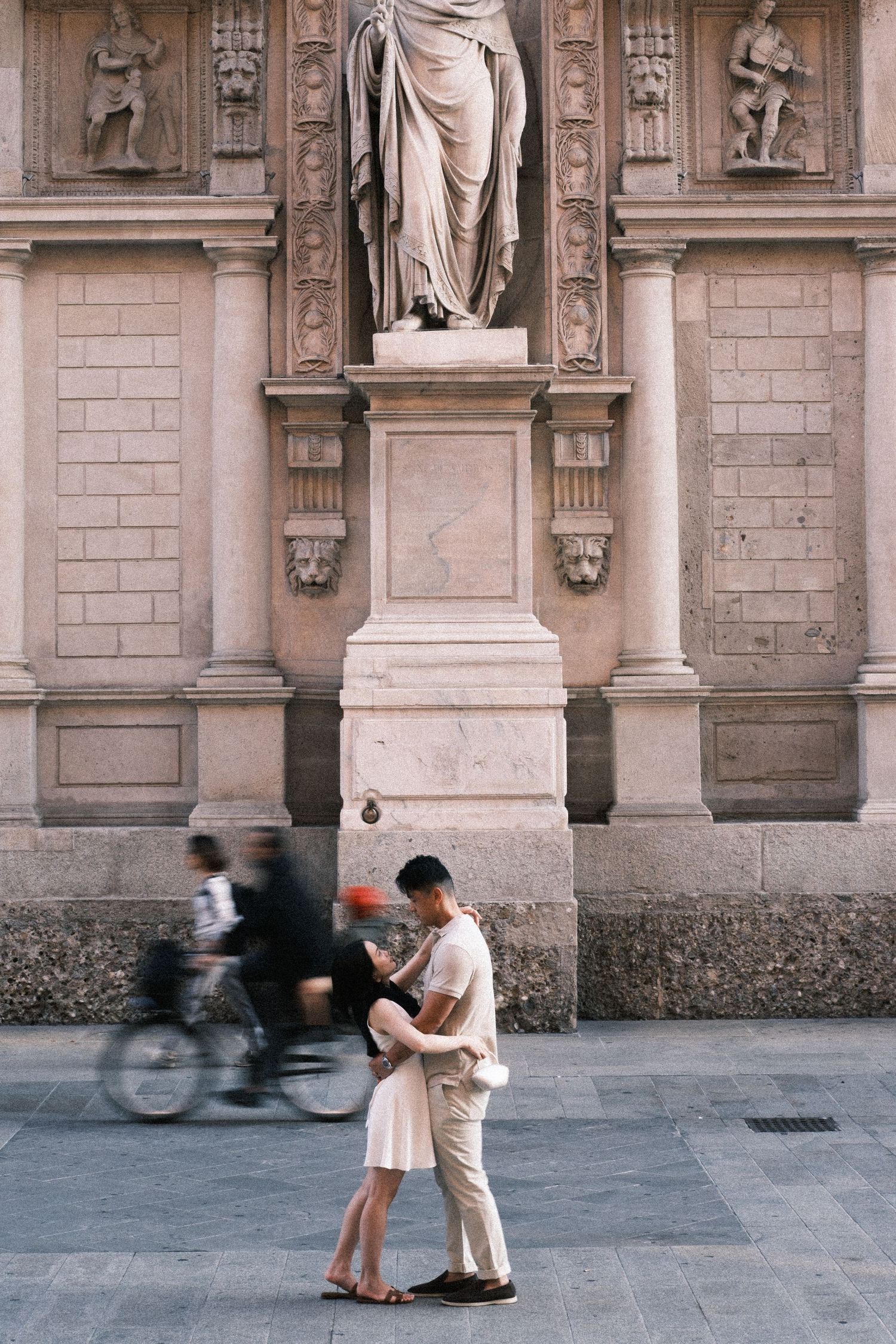 Couple embracing in front of a classical statue and ornate building facade, with a blurred cyclist passing by.