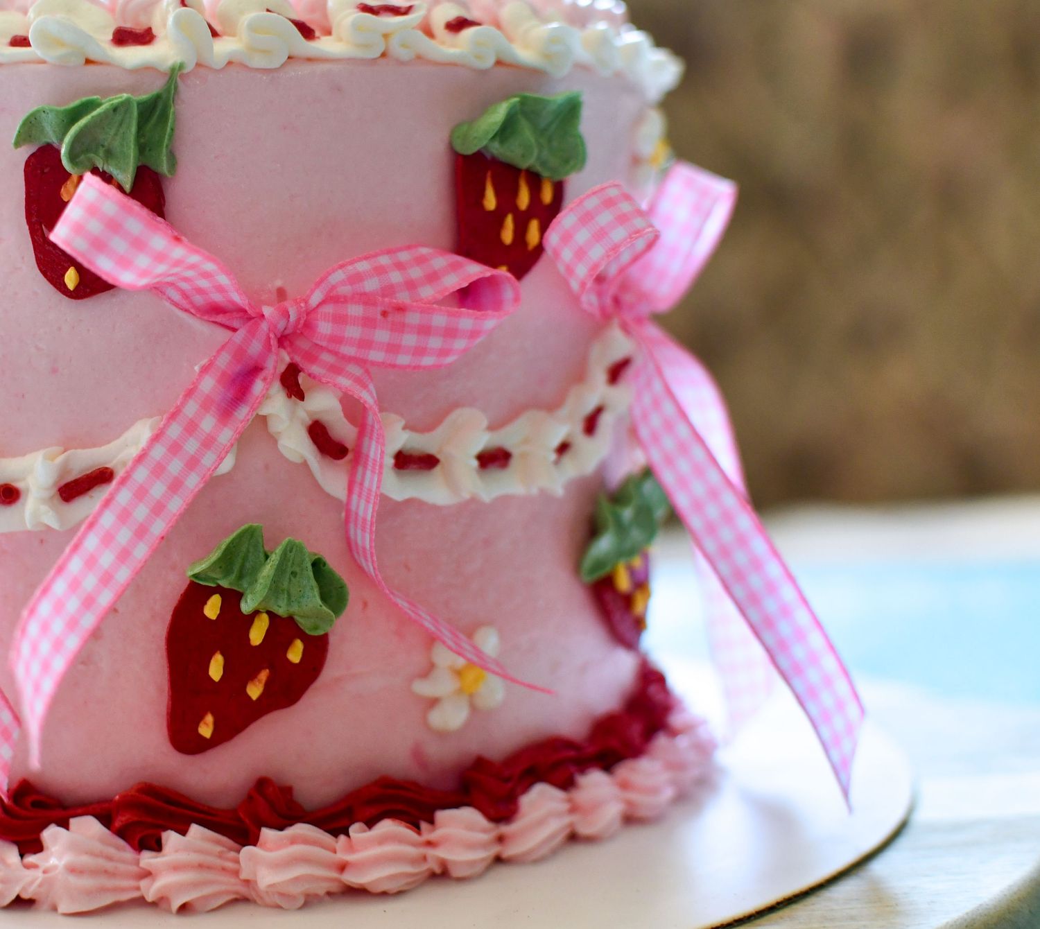 Pink two-tier strawberry cake with gingham ribbon bows, white lace frosting, and red strawberry decorations on a white stand.