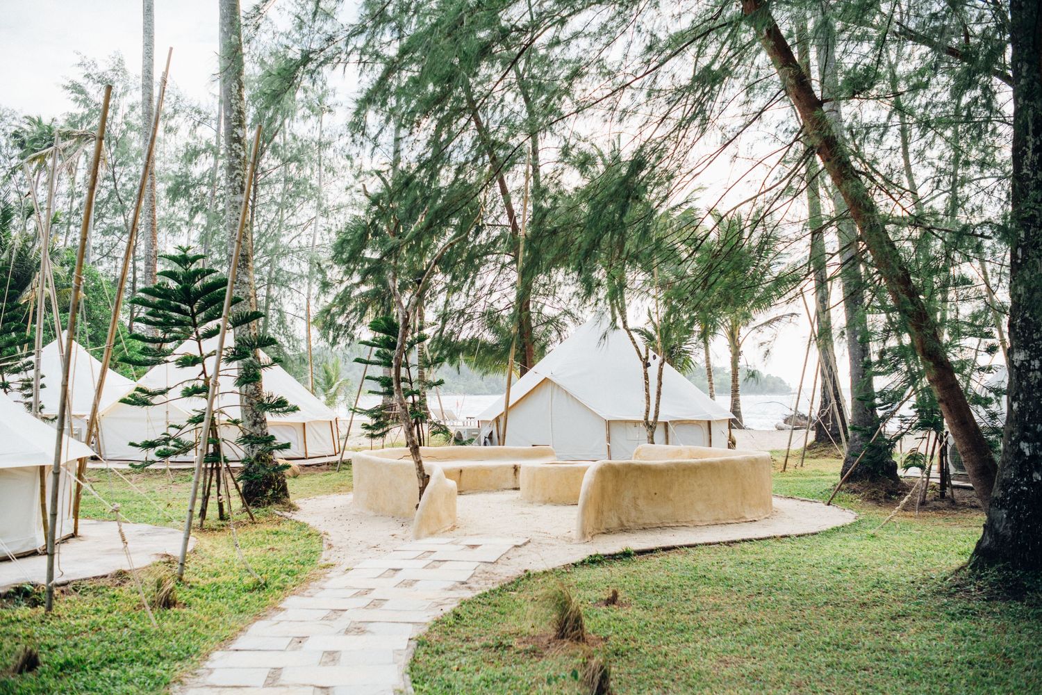Glamping tents nestled among tall trees on a tropical beach resort with stone pathways and lush green grass.