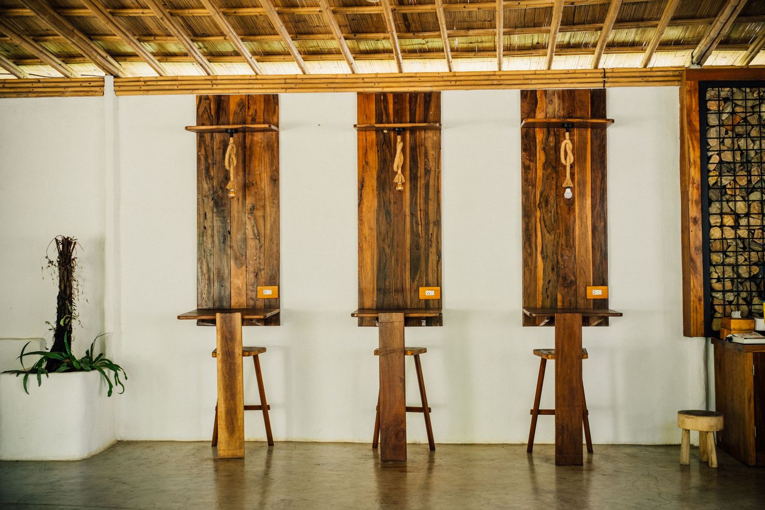 Three rustic wooden bar stools with rope details stand against a white wall under a bamboo ceiling.