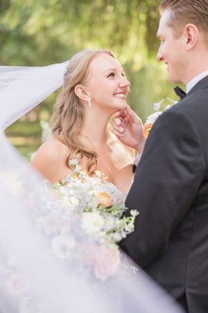 Bride smiling up at groom during outdoor wedding ceremony, holding pastel floral bouquet with veil flowing in breeze.