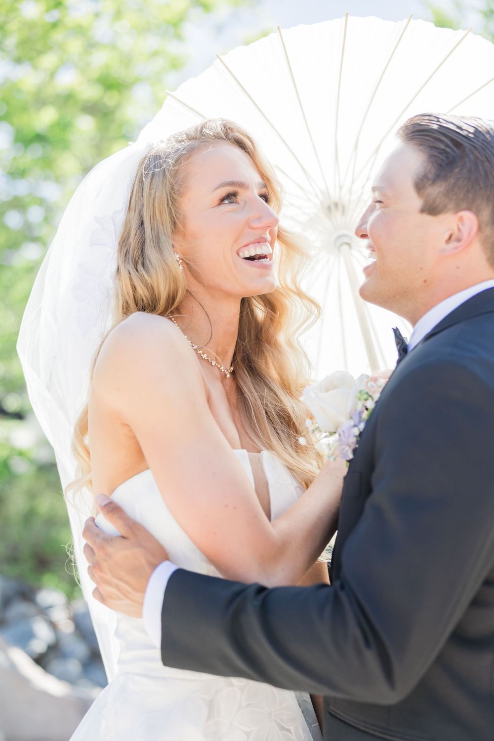 Bride and groom laughing together under a white parasol on their wedding day.