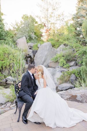 A bride and groom share a romantic kiss while seated on a bench surrounded by lush greenery and rocks on their wedding day.