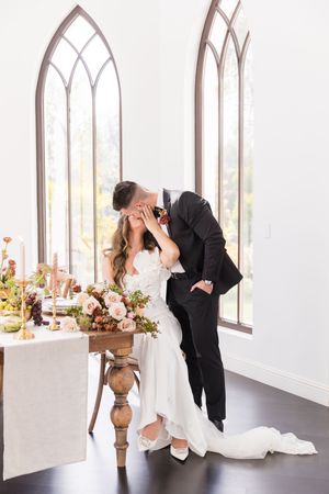 Bride and groom share a romantic kiss at elegantly decorated wedding table with floral arrangements in a bright chapel.