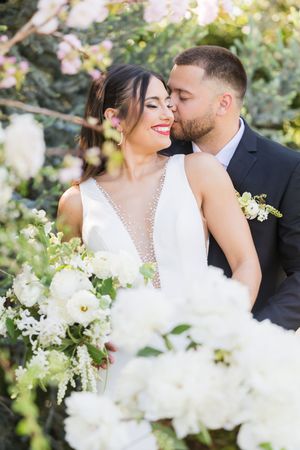 Groom in black suit kisses bride in white gown surrounded by lush white florals and pink blossoms outdoors.