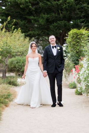 Bride in white strapless gown and groom in black tuxedo hold hands walking along a garden path on their wedding day.