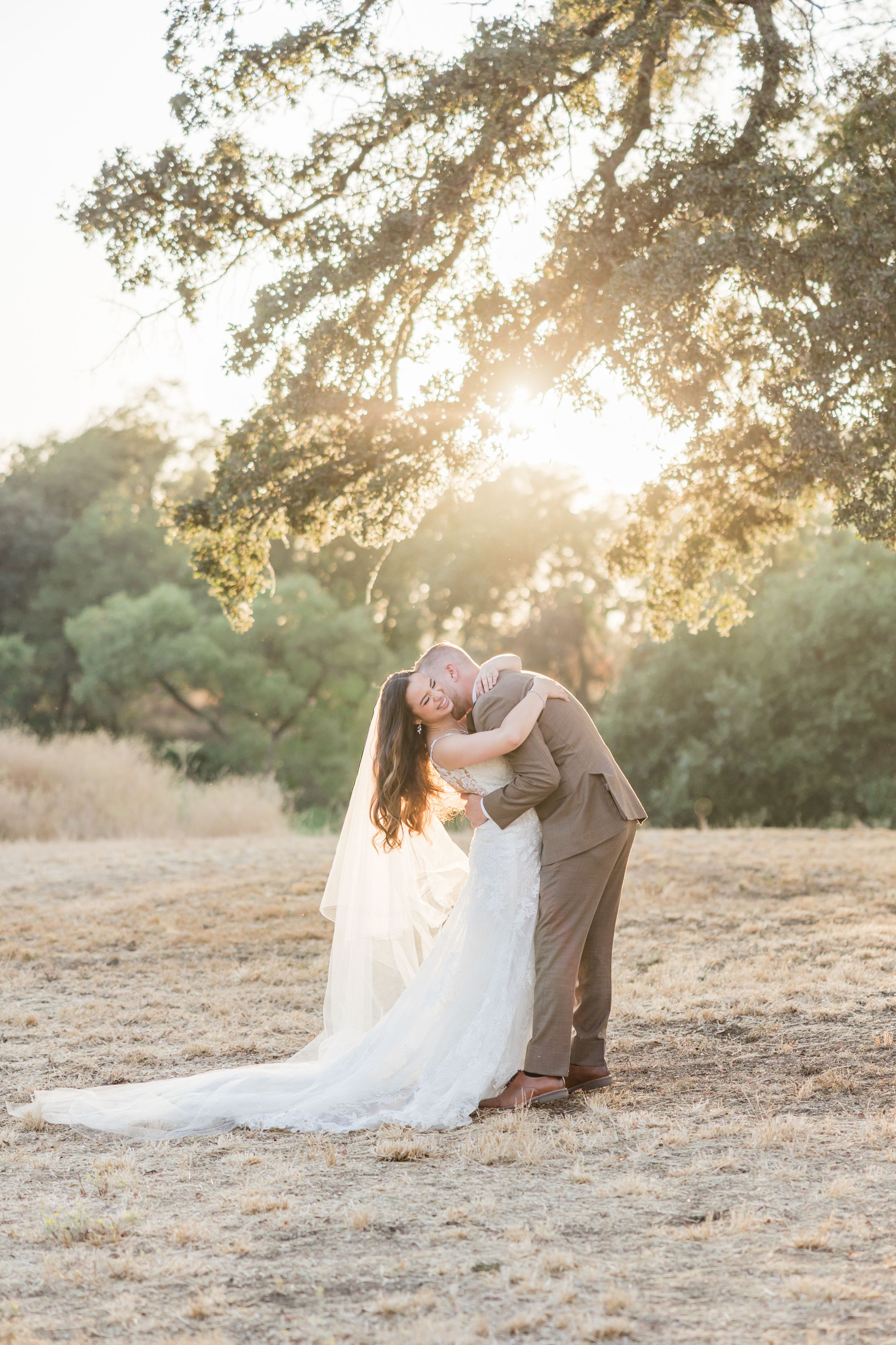 Bride and groom share a romantic embrace in golden sunset light beneath oak trees in a dry meadow.