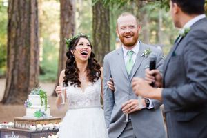 Laughing bride and groom stand together outdoors at rustic woodland wedding reception with cake table.