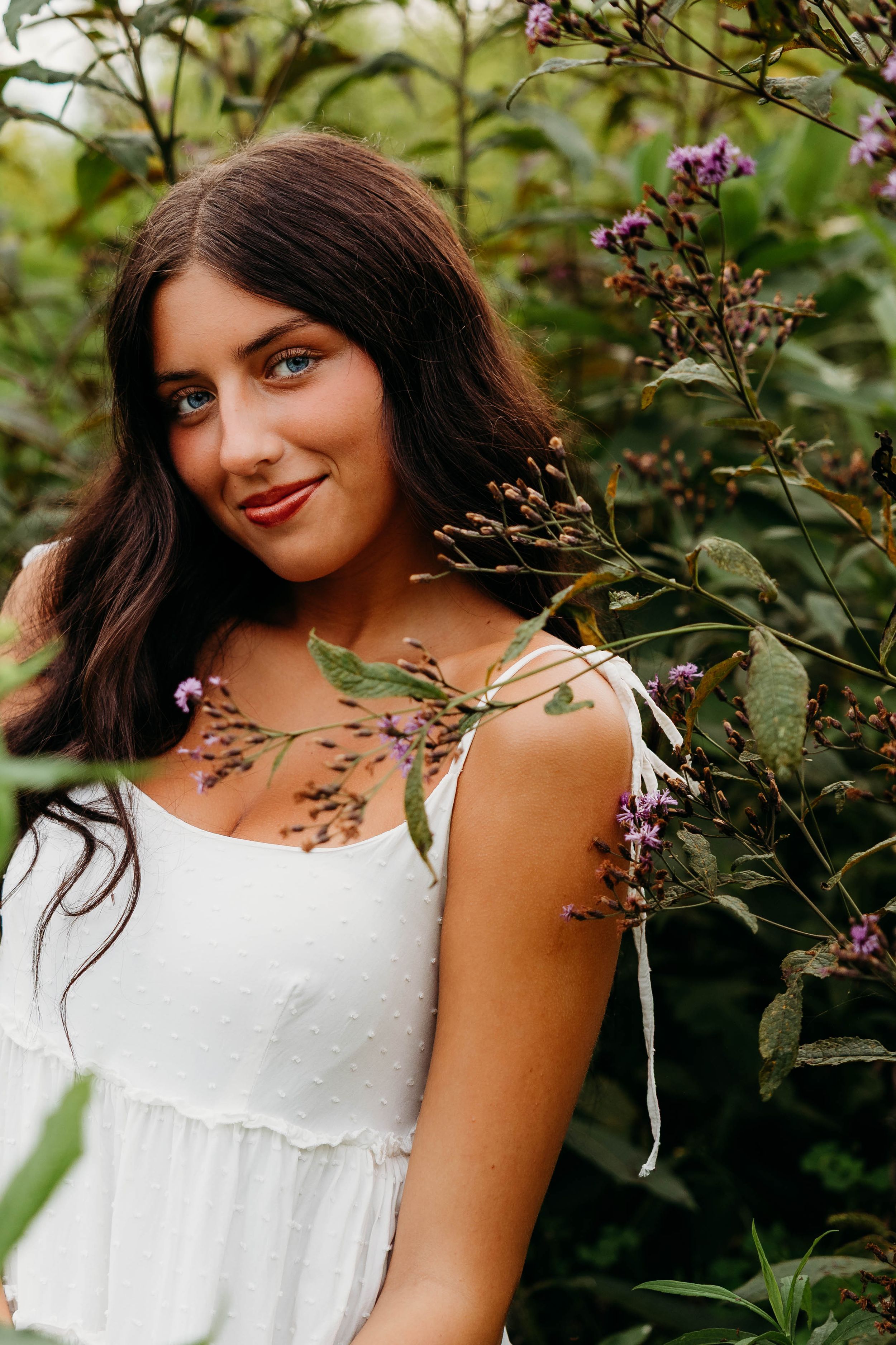 Young brunette woman in white sundress poses among purple wildflowers and green foliage outdoors.