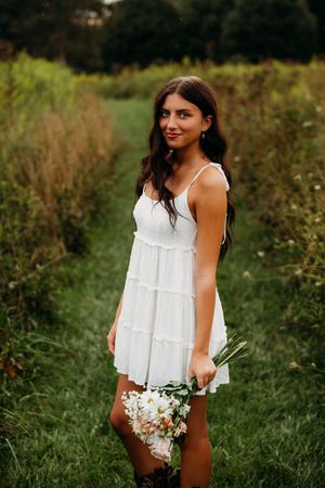 Young woman in white sundress holding wildflowers, standing in a lush green field path, smiling softly.