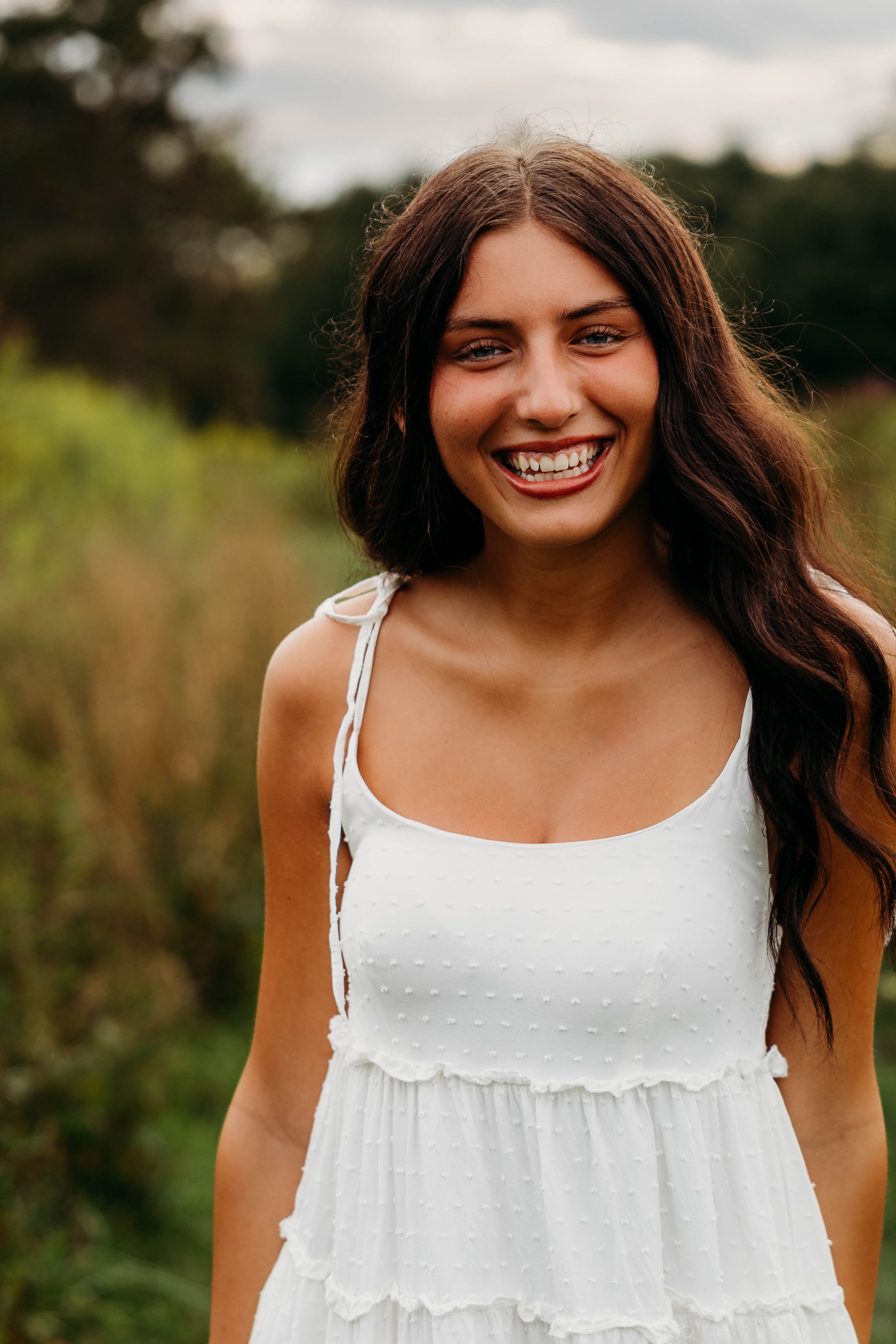 Smiling young woman with long brown hair wearing a white sundress outdoors in a green field.