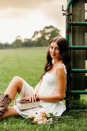 Young woman in white dress and cowboy boots sits by rustic green gate in field, holding a wooden box with flowers nearby.