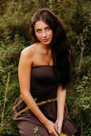 Young woman with long dark hair and blue eyes wearing a brown strapless dress, sitting outdoors among green foliage.