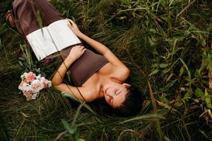 Woman lying in green grass with pink flowers, wearing brown top and white skirt, eyes closed, serene outdoor portrait.