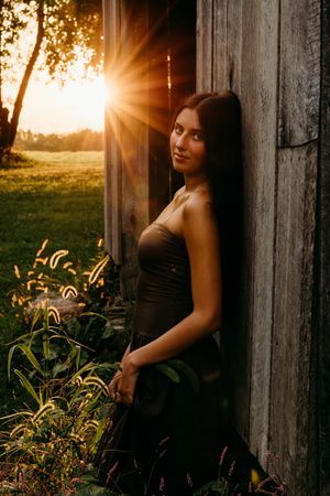 Young woman in dark dress leaning against rustic wooden barn wall at golden sunset hour.