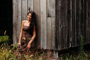 Young woman in brown strapless dress leaning against rustic wooden barn wall surrounded by wild greenery.