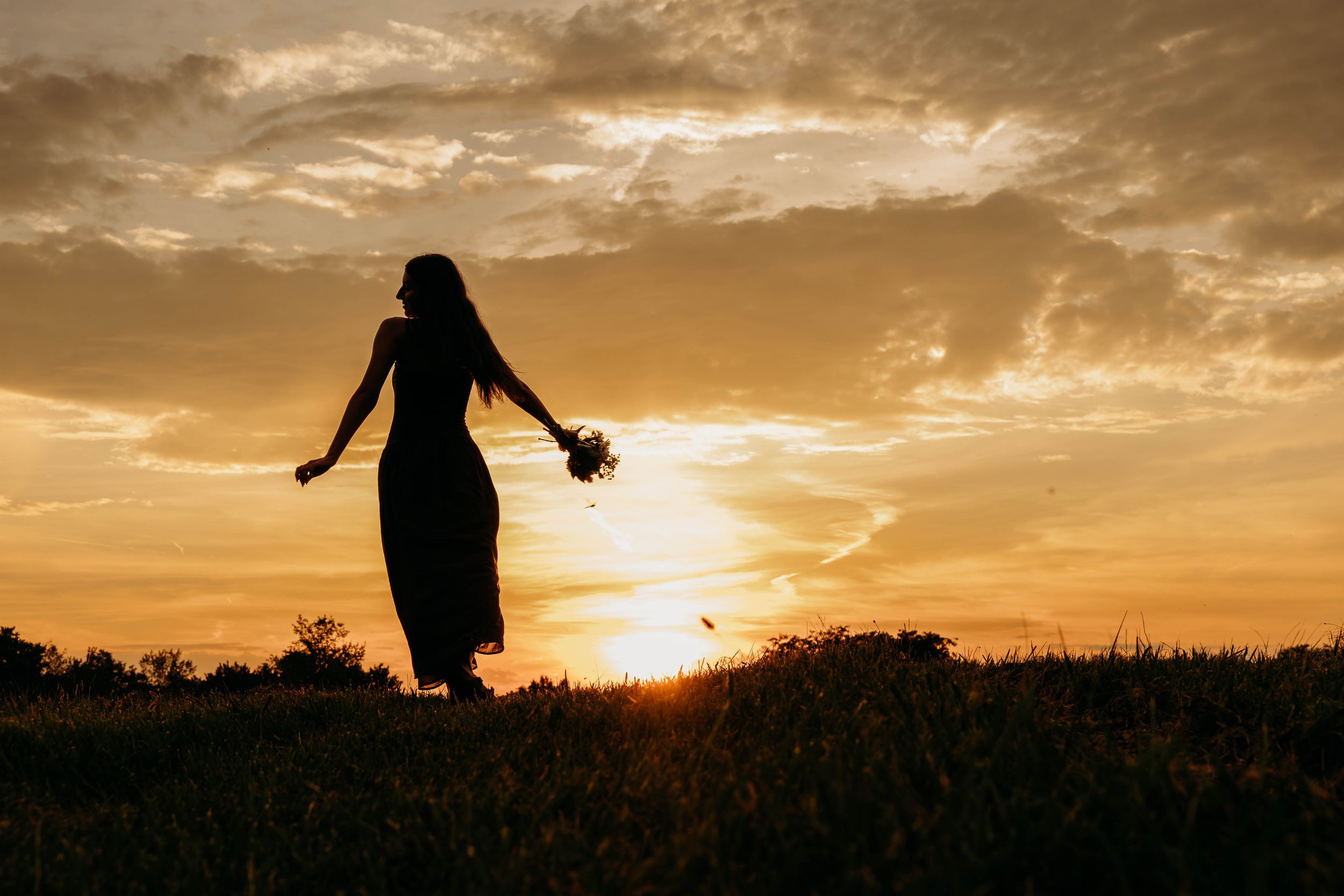 Silhouette of a woman holding flowers standing in a golden sunset field with dramatic cloudy sky.
