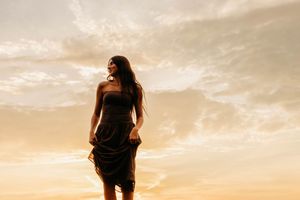 Woman in black strapless dress standing against golden sunset sky with dramatic clouds.