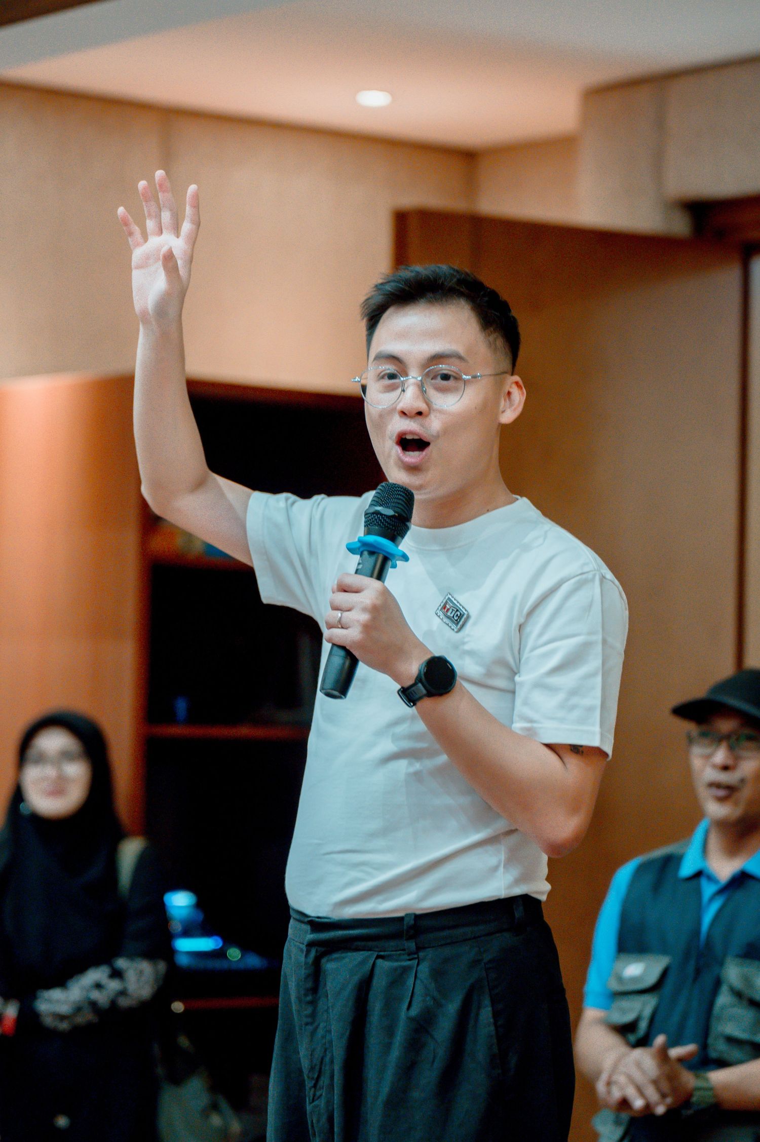 A young man in glasses and white t-shirt speaks into a microphone with hand raised at an indoor event.