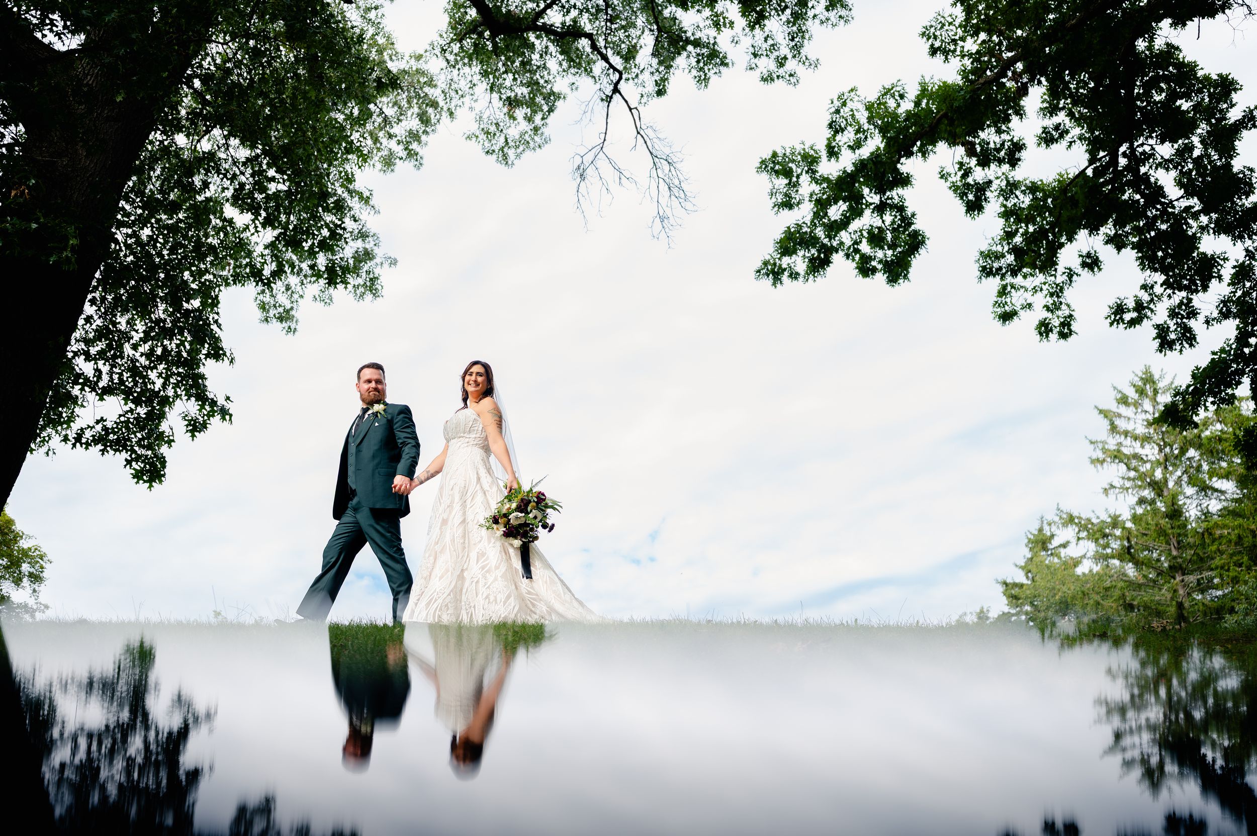 Bride and groom walk together outdoors, reflected in water below, framed by lush green trees on their wedding day.