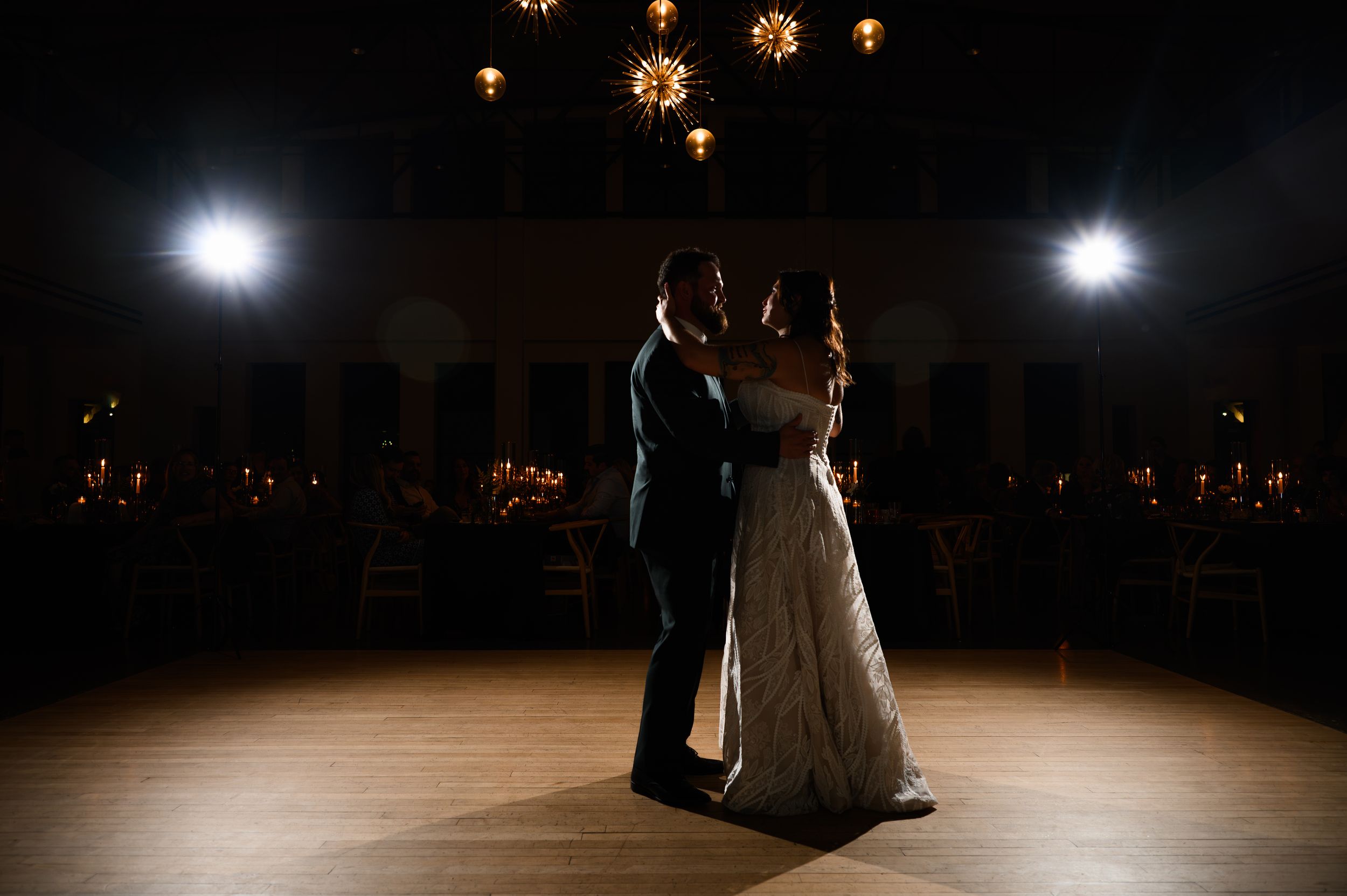 Bride and groom share first dance in spotlight at elegant wedding reception venue with candlelit tables.