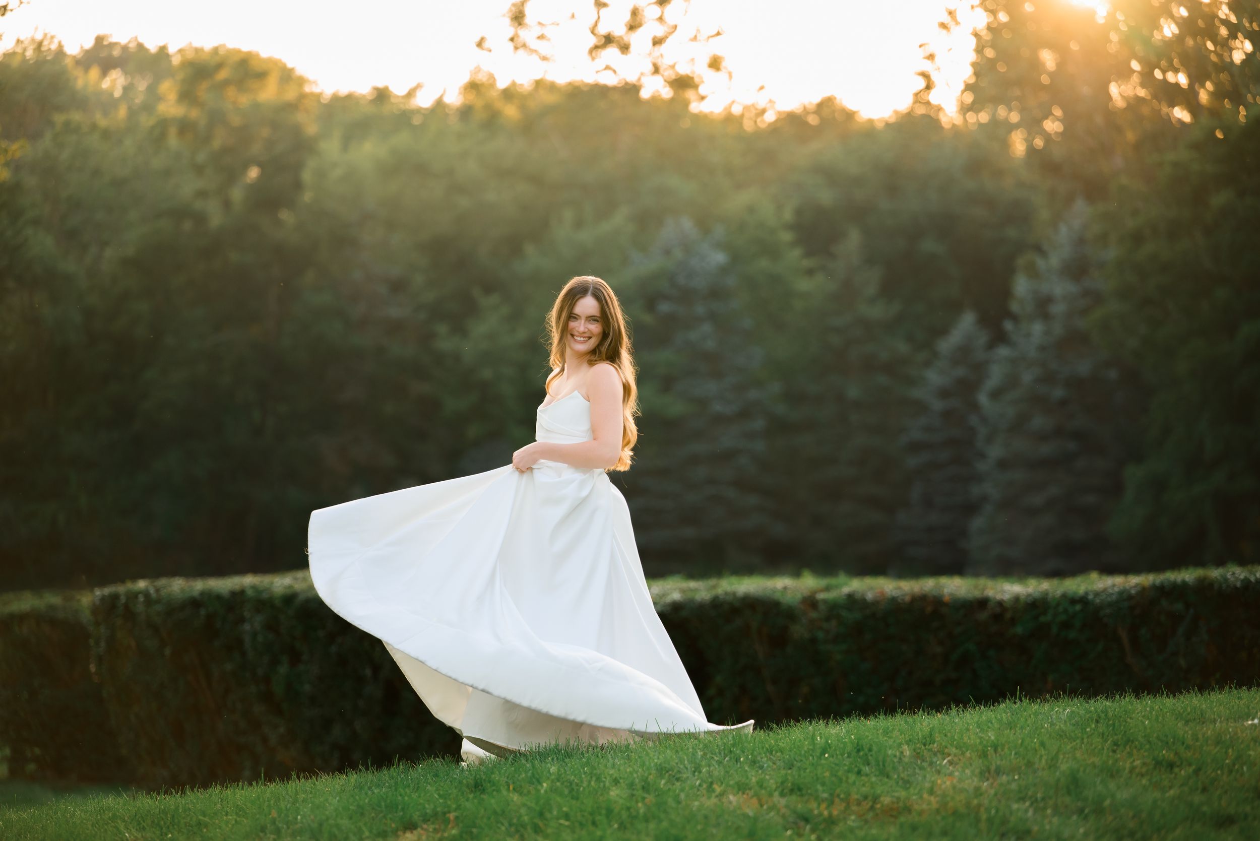 Bride in white flowing gown twirling in sunlit garden with lush green hedges and trees at golden hour.