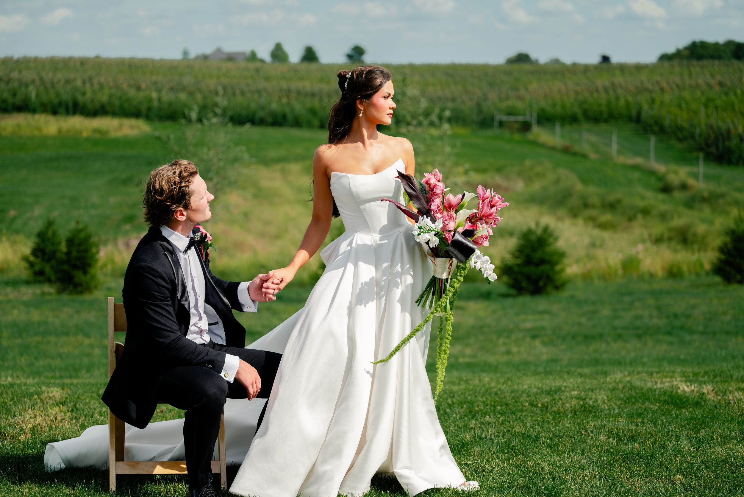 Bride in white gown holds bouquet while groom sits outdoors on lush green farm wedding venue.
