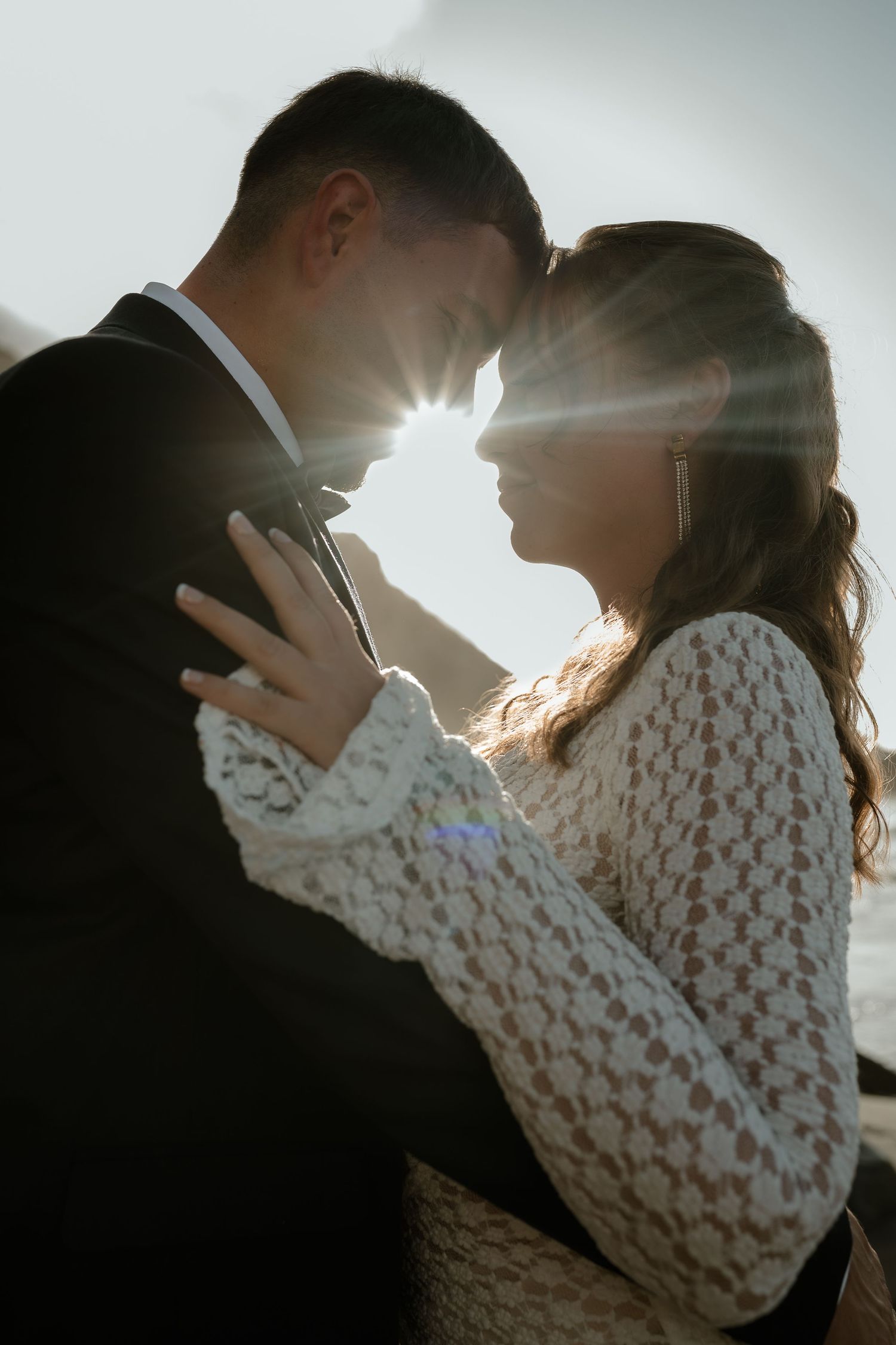 Couple in wedding attire sharing a romantic moment with sun flare between them outdoors.