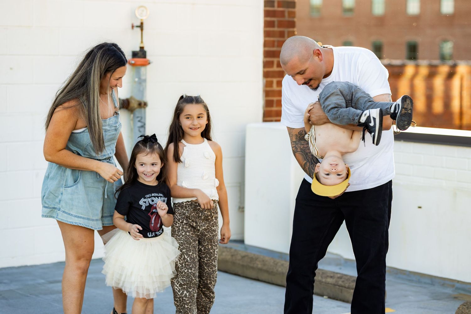 A tattooed man playfully holds a baby upside down while a woman and two girls smile outdoors.