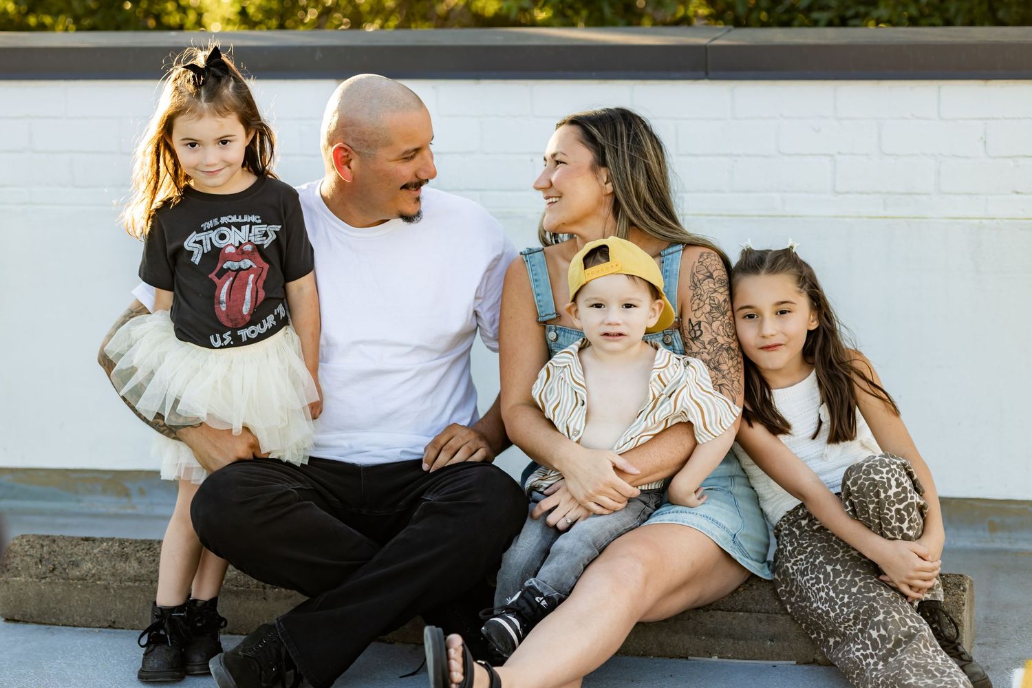 Happy family of five sitting outdoors on steps, smiling together in casual summer outfits against a white brick wall.