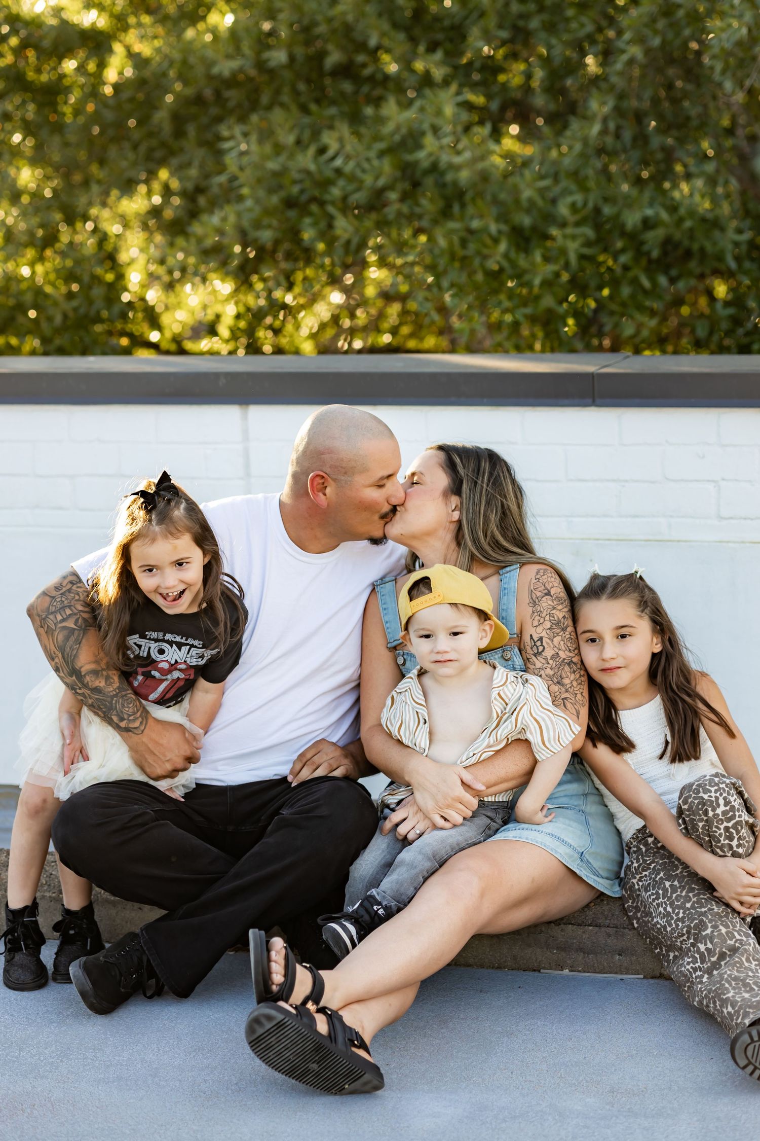 A tattooed couple kisses while sitting with their three young children outdoors against a white brick wall.