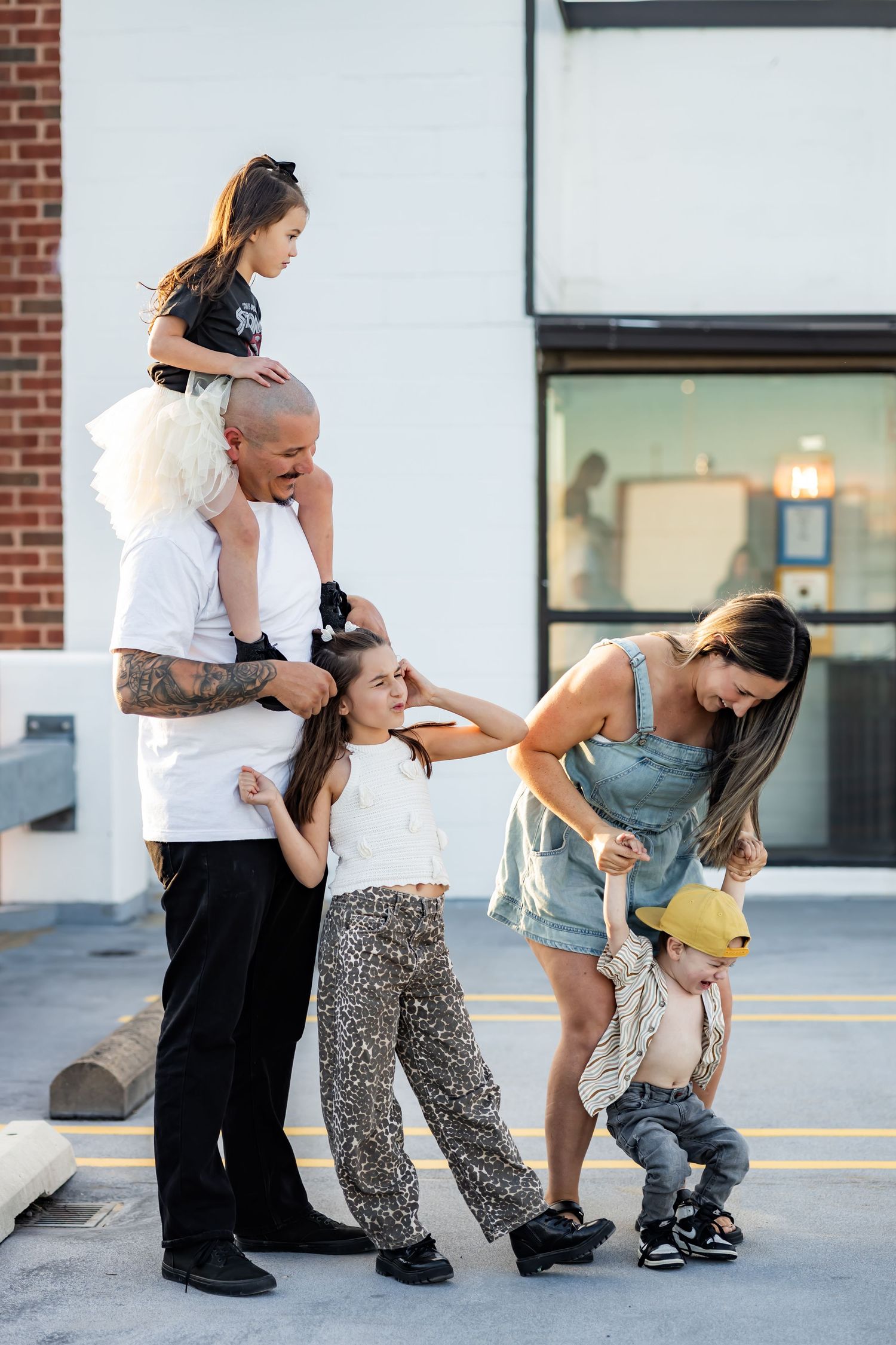 A tattooed man holds a child on his shoulders while a woman tends to a toddler, surrounded by two girls outdoors.