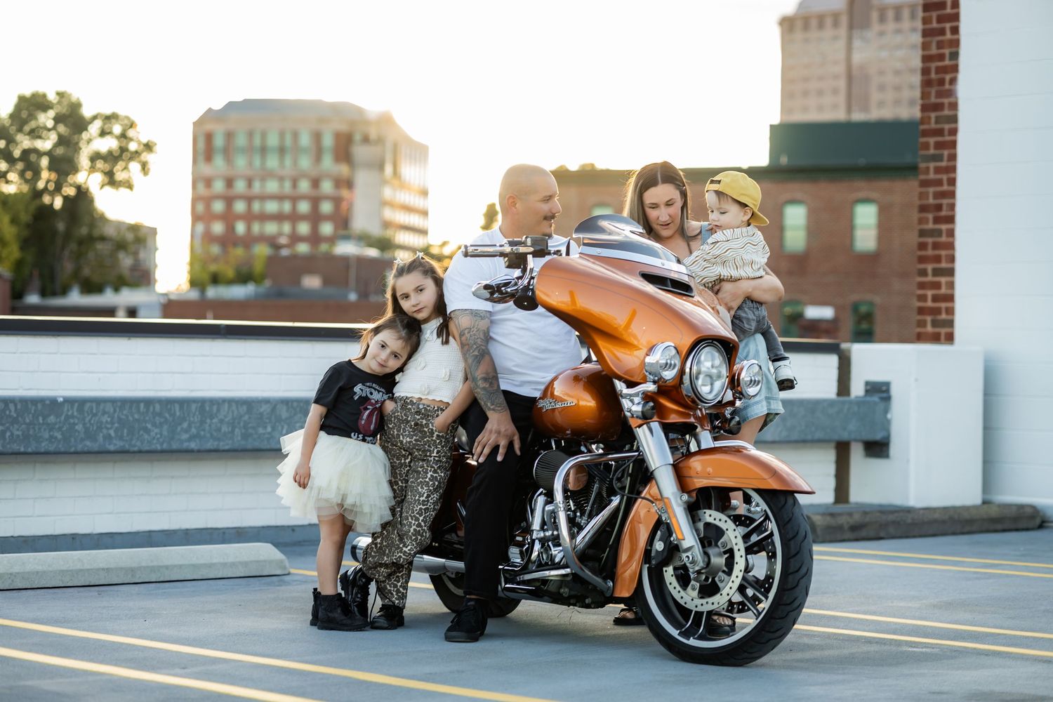Family posing with orange Harley-Davidson motorcycle on rooftop parking lot at sunset with city buildings in background.