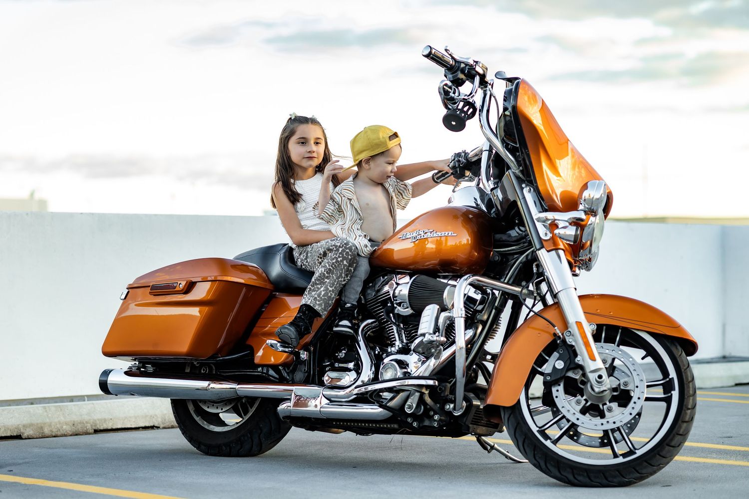 Two children sitting on a shiny orange Harley-Davidson motorcycle in a parking lot.