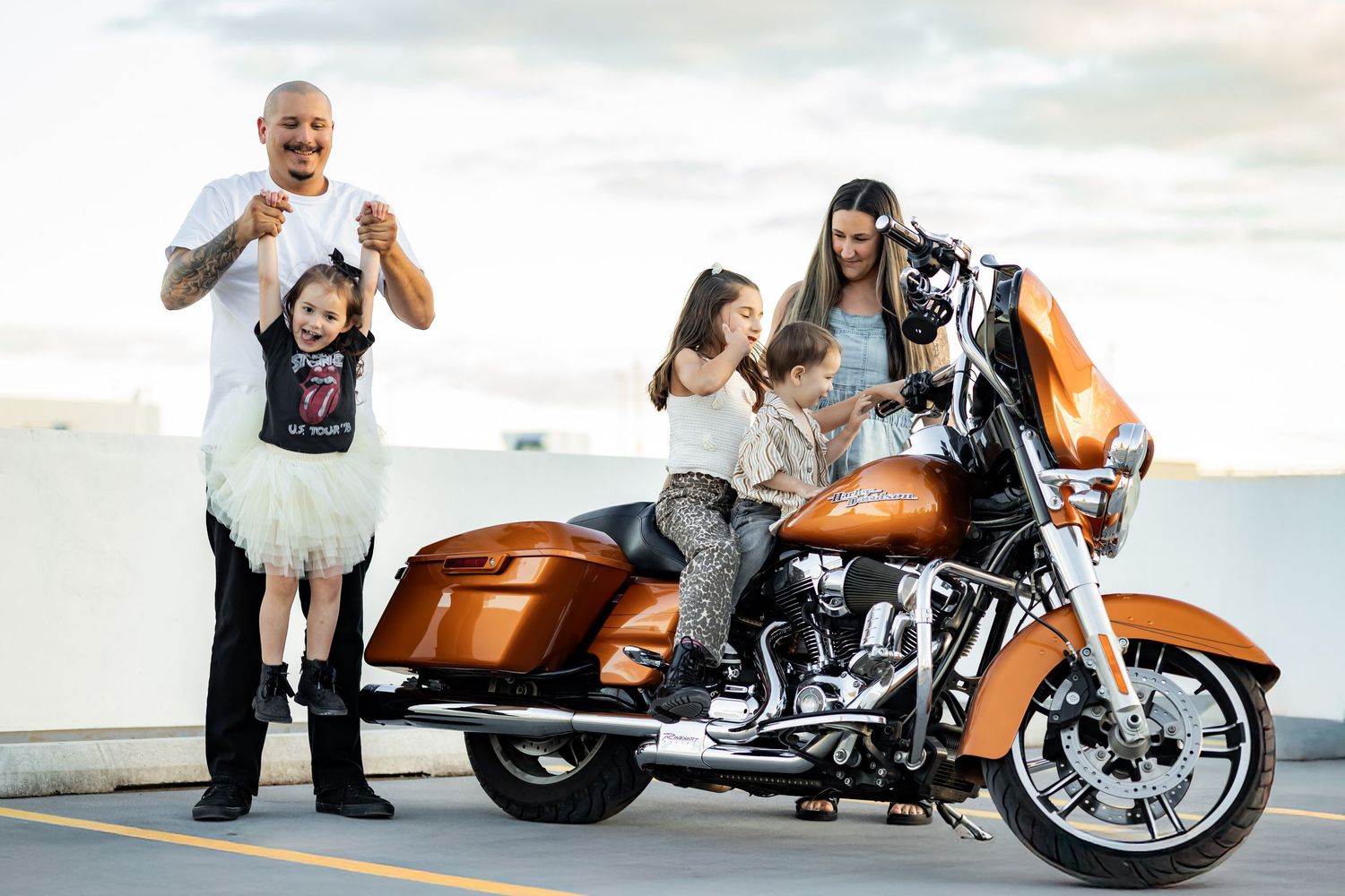 Family posing with an orange Harley-Davidson motorcycle outdoors, including a tattooed man holding a child.