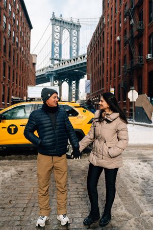 Couple holding hands in DUMBO Brooklyn with Manhattan Bridge and yellow NYC taxi cab in snowy winter backdrop.