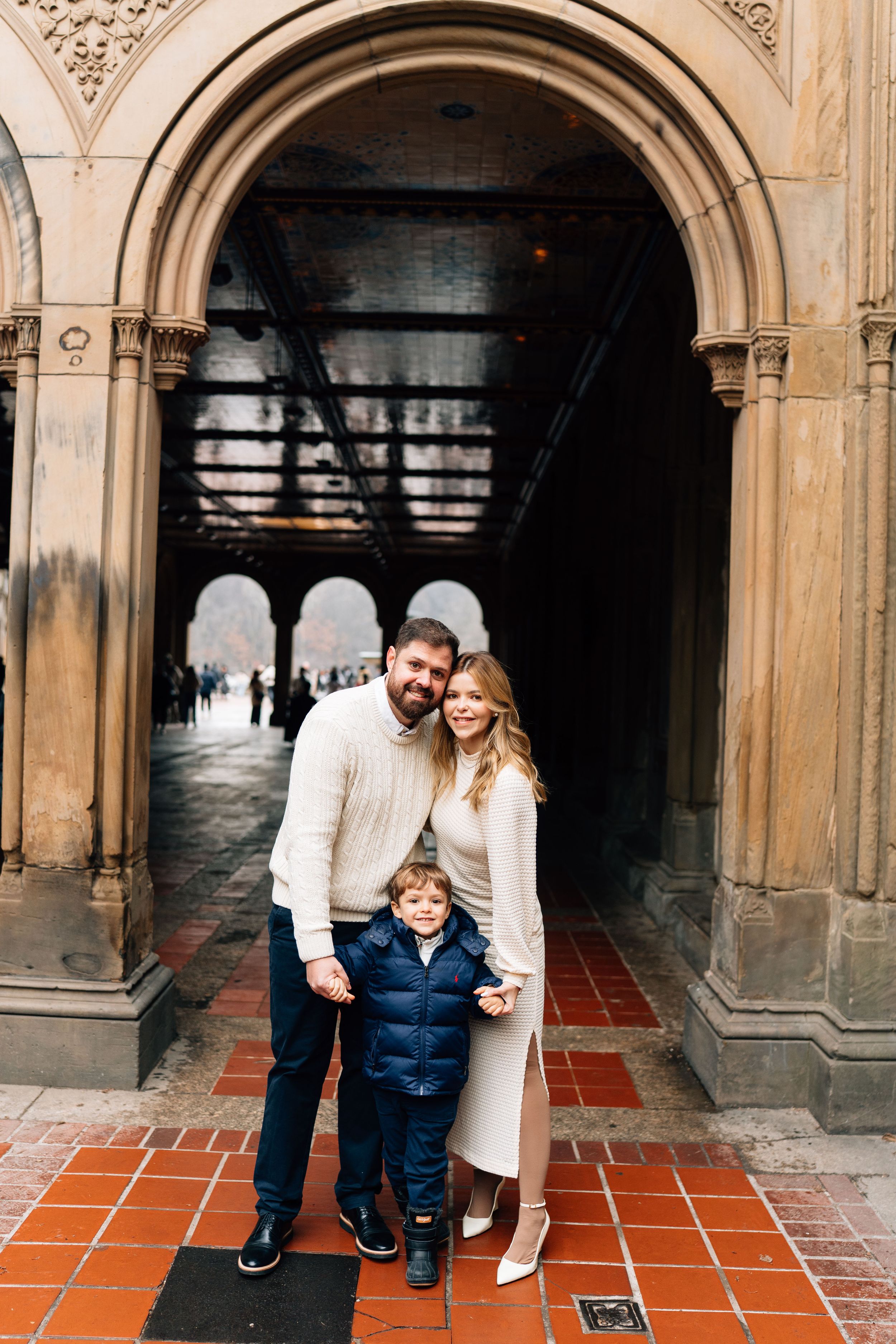Family of three posing under ornate stone arch at Bethesda Terrace in Central Park, NYC.