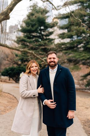 Smiling couple in winter coats posing together in a park with evergreen trees and city buildings in background.