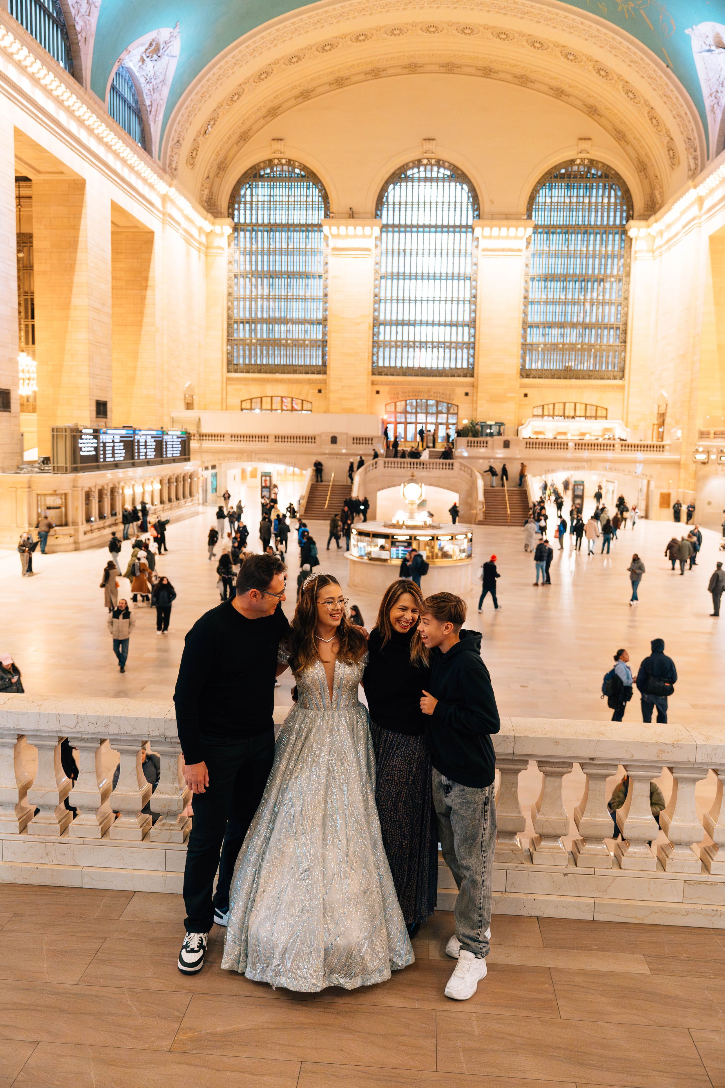 Family posing in elegant attire at Grand Central Terminal NYC, woman in silver ball gown with arched windows behind.
