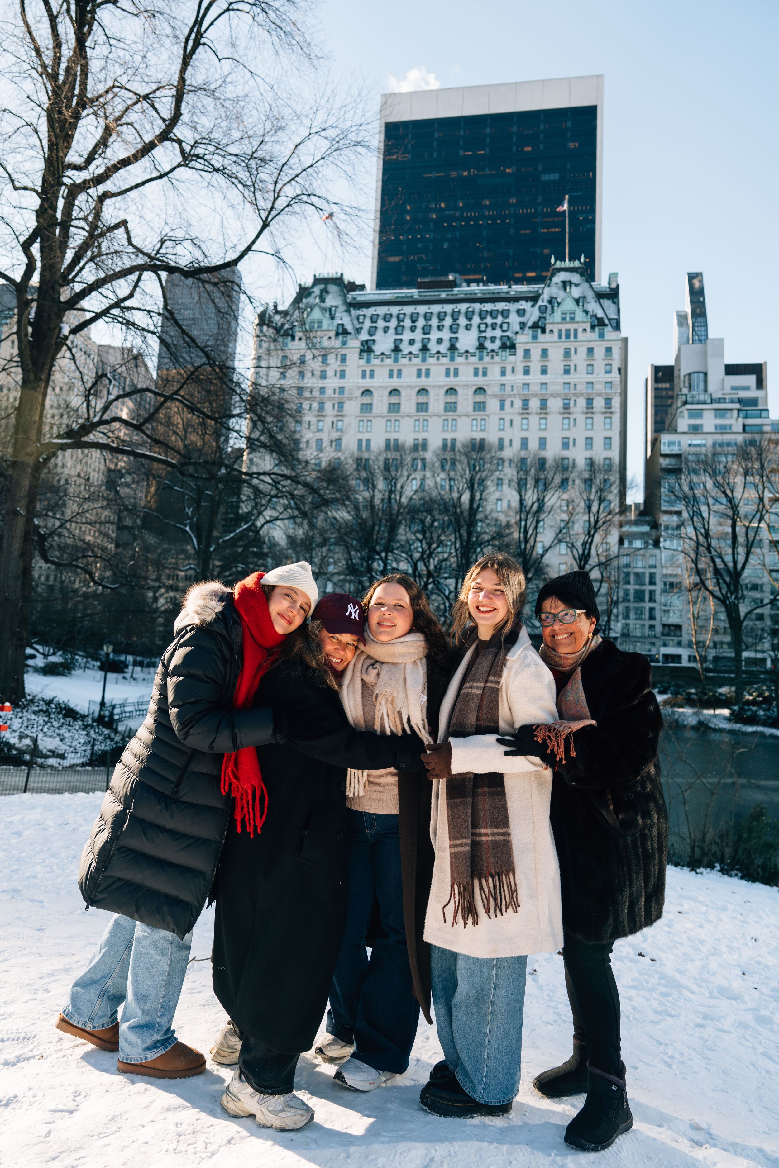 Five women pose together smiling in snowy Central Park NYC with The Plaza Hotel visible in the background.
